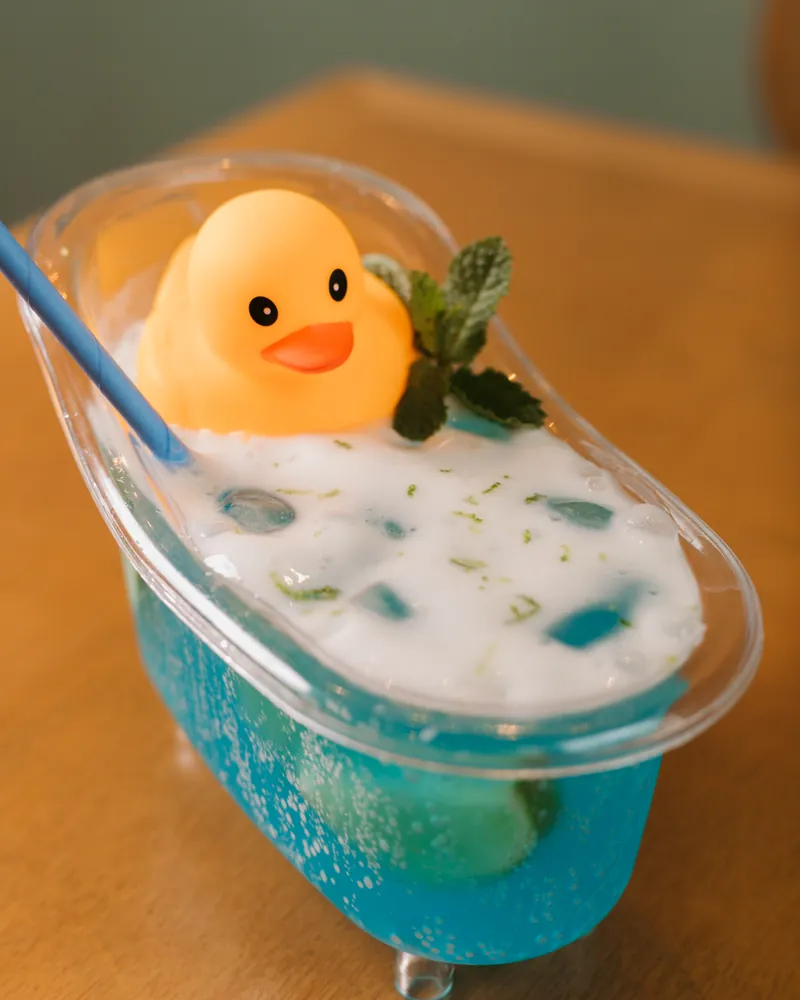 Drink azul servido em uma pequena banheira transparente com patinho amarelo e hortelã