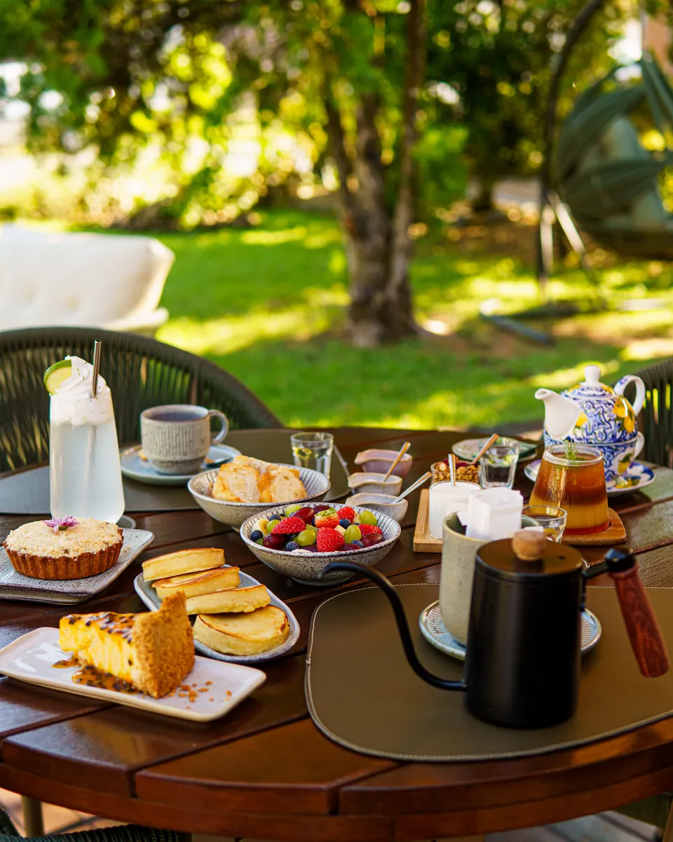 Mesa posta no jardim do Momentos, com chá, bolo, torradas, frutas e cadeiras trançadas verdes sob as árvores
