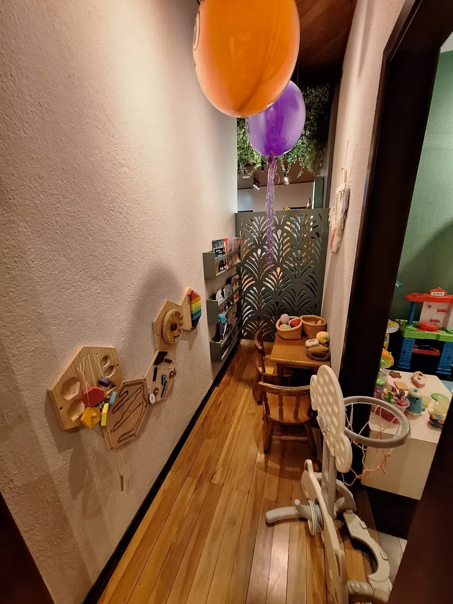 Corredor ao lado do Espaço Kids, com painéis de brinquedos de madeira na parede e balões coloridos